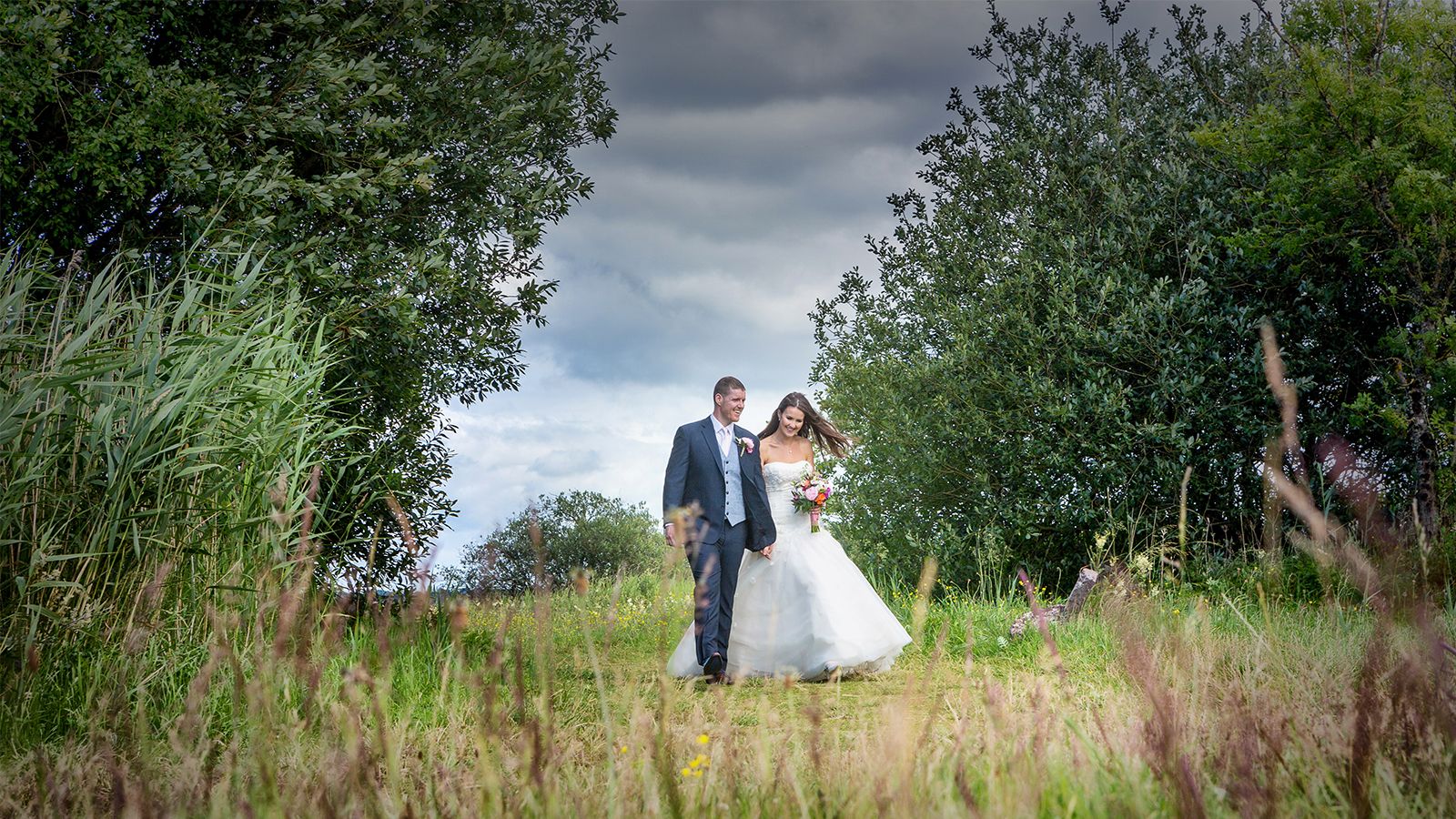 A wedding photographer captures a bride and groom walking through tall grass.