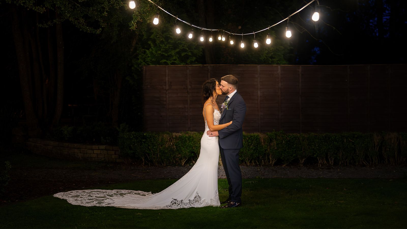 A newlywed couple kissing under string lights at night in a garden, surrounded by trees, with the bride in a long white gown and the groom in a suit.
