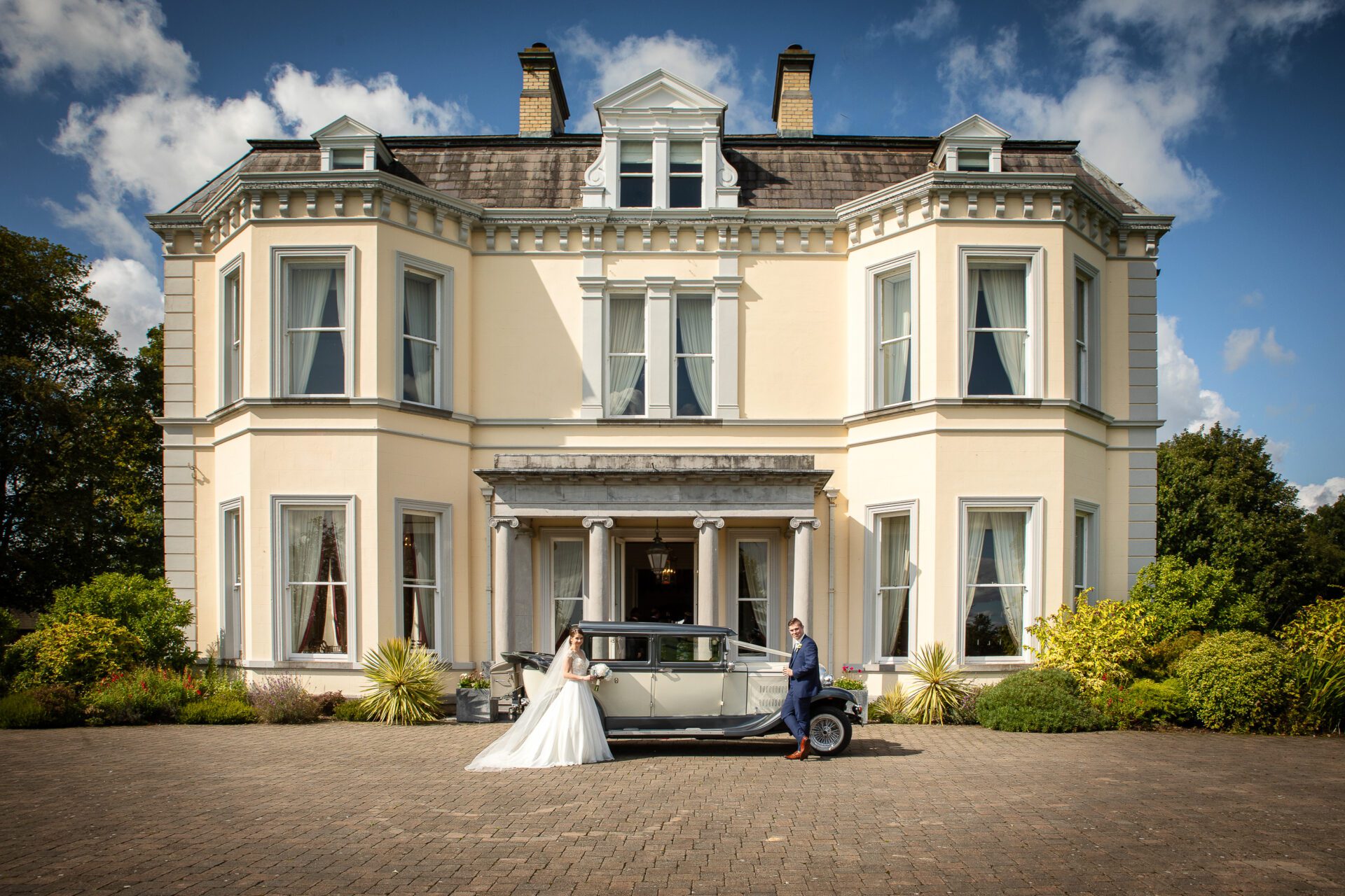 A bride and groom stand by a vintage car in front of an elegant, large house with lush greenery under a clear blue sky. the groom is holding the car door open for the bride.