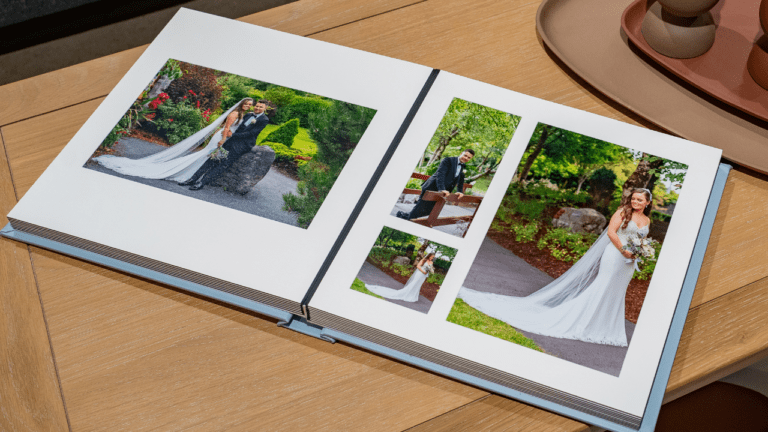 A wedding album open on a wooden table, displaying photos of a bride and groom in a garden setting. The left page shows a couple's portrait and the right page features multiple images of the bride in her dress. In the background is a tray with cups.