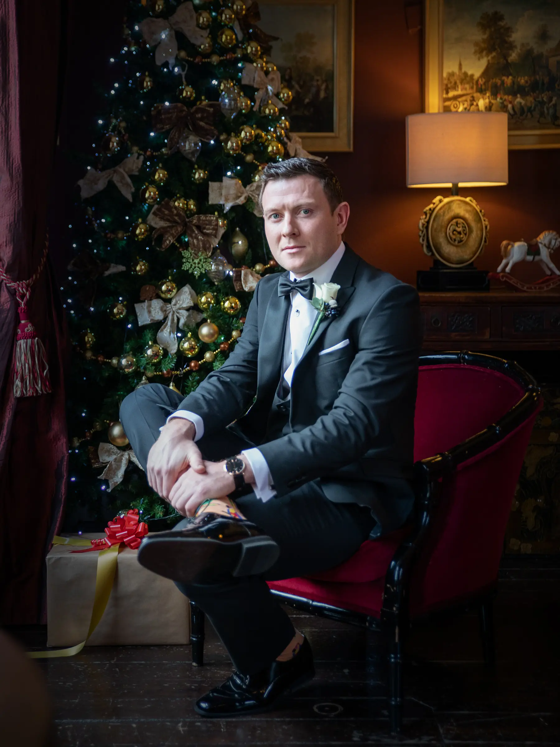 An elegant groom in a tuxedo relaxing next to a beautifully decorated Christmas tree. Read our privacy policy for information on how we handle your personal data.