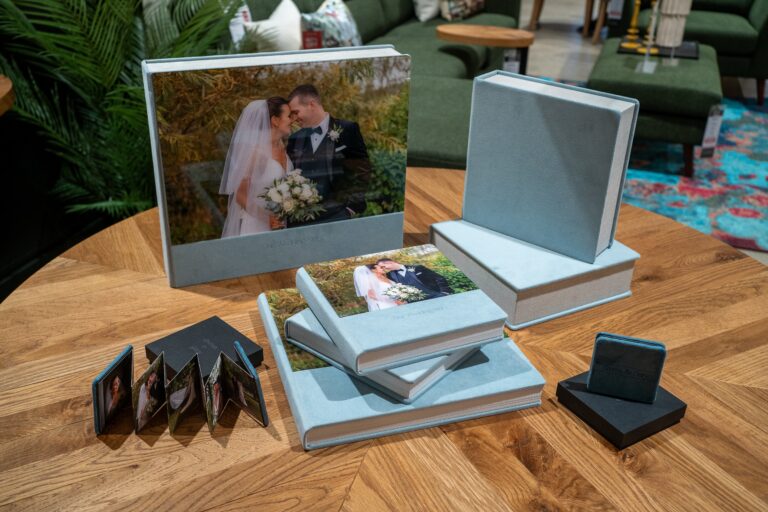 A display of wedding photography packages on a wooden table, including a framed photo, open photo books, and small accordion-style mini albums, all featuring images of a bride and groom.